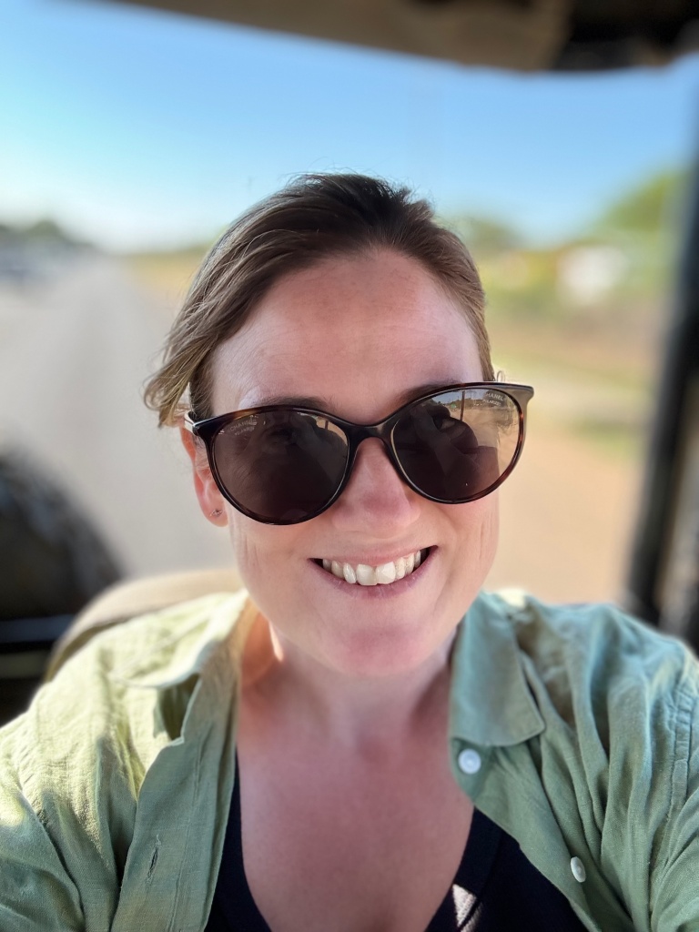 Picture of me (Sharon O'Dea) in a safari truck on the Botswana-Zimbabwe border. I am wearing sunglasses and a khaki green shirt. I am smiling and look happy. Because I am.
