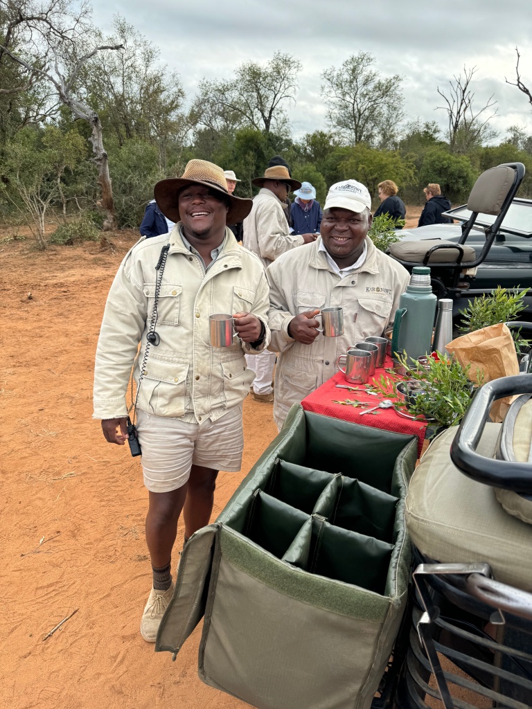 Two safari guides, standing next to a safari truck. They are holding mugs and smiling.