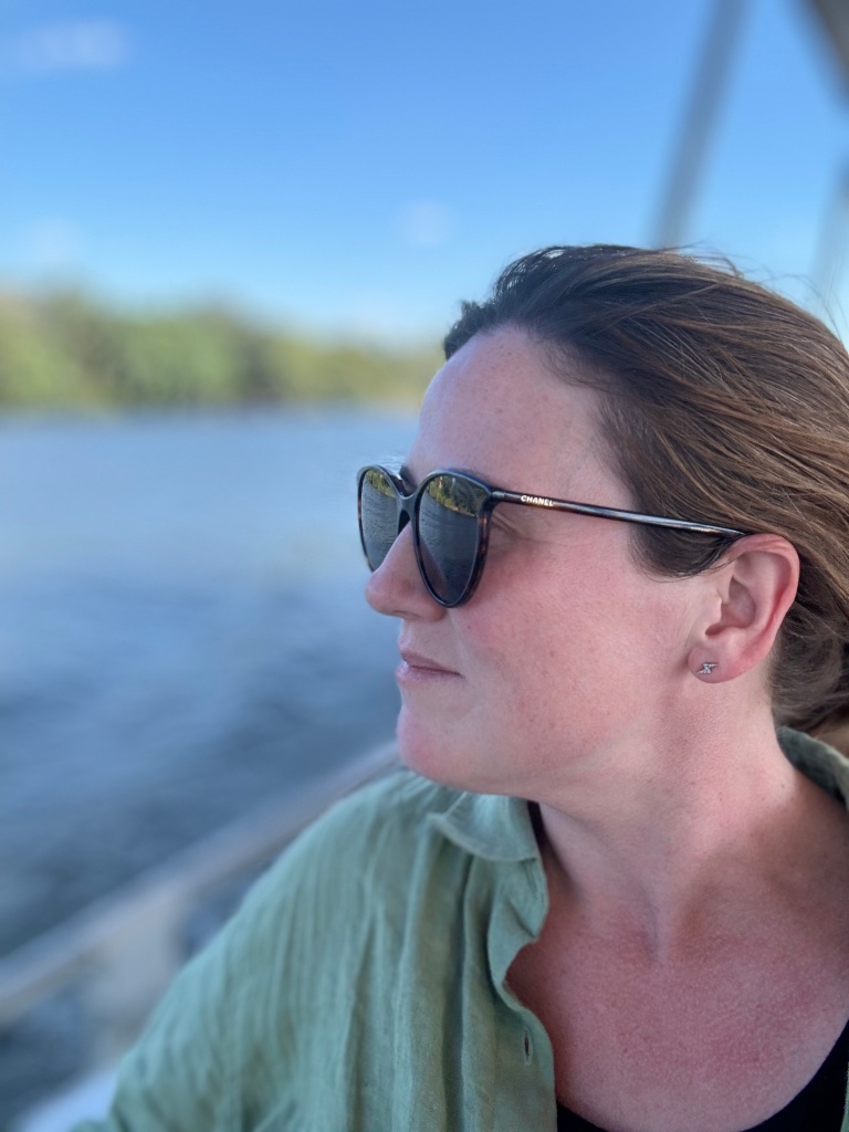 A photo of me looking into the distance from a boat on the Chobe river, Botswana. I am wearing sunglasses and a green shirt.